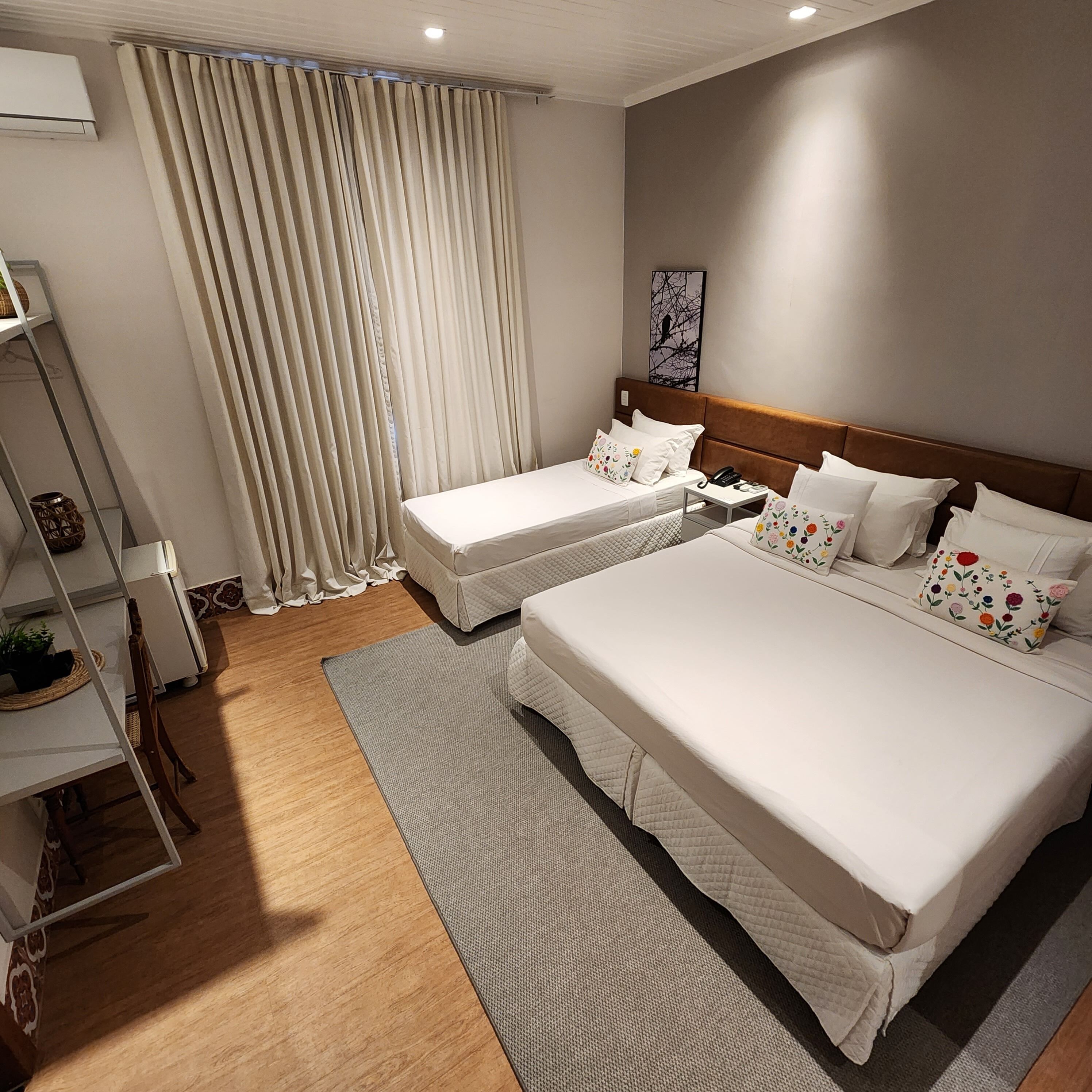 A hotel room with two beds, a small table, a chair, a shelf with decorative items, and a window with curtains.
