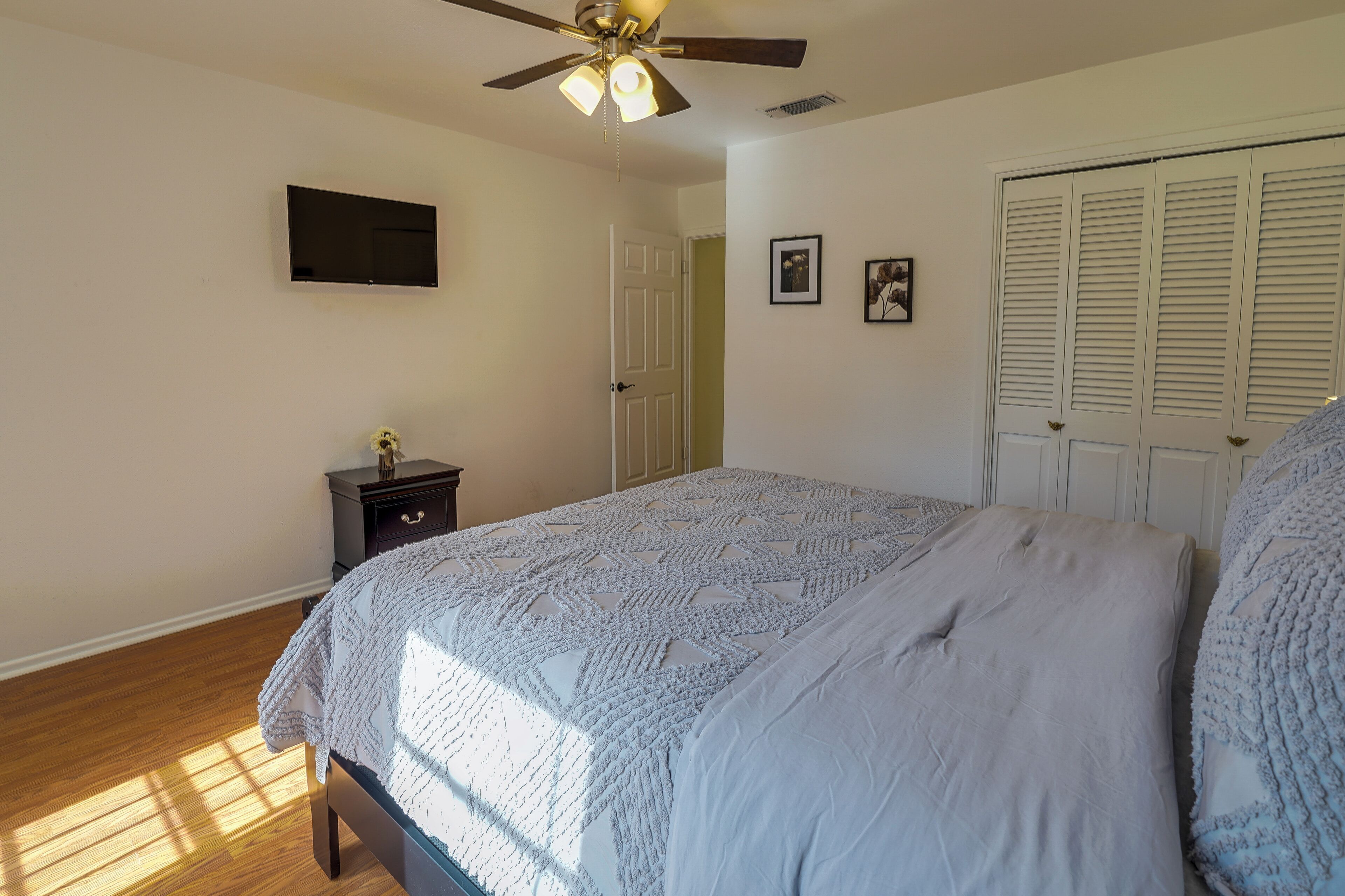 A bedroom with a bed, a ceiling fan, a TV mounted on the wall, and a door with louvered shutters.