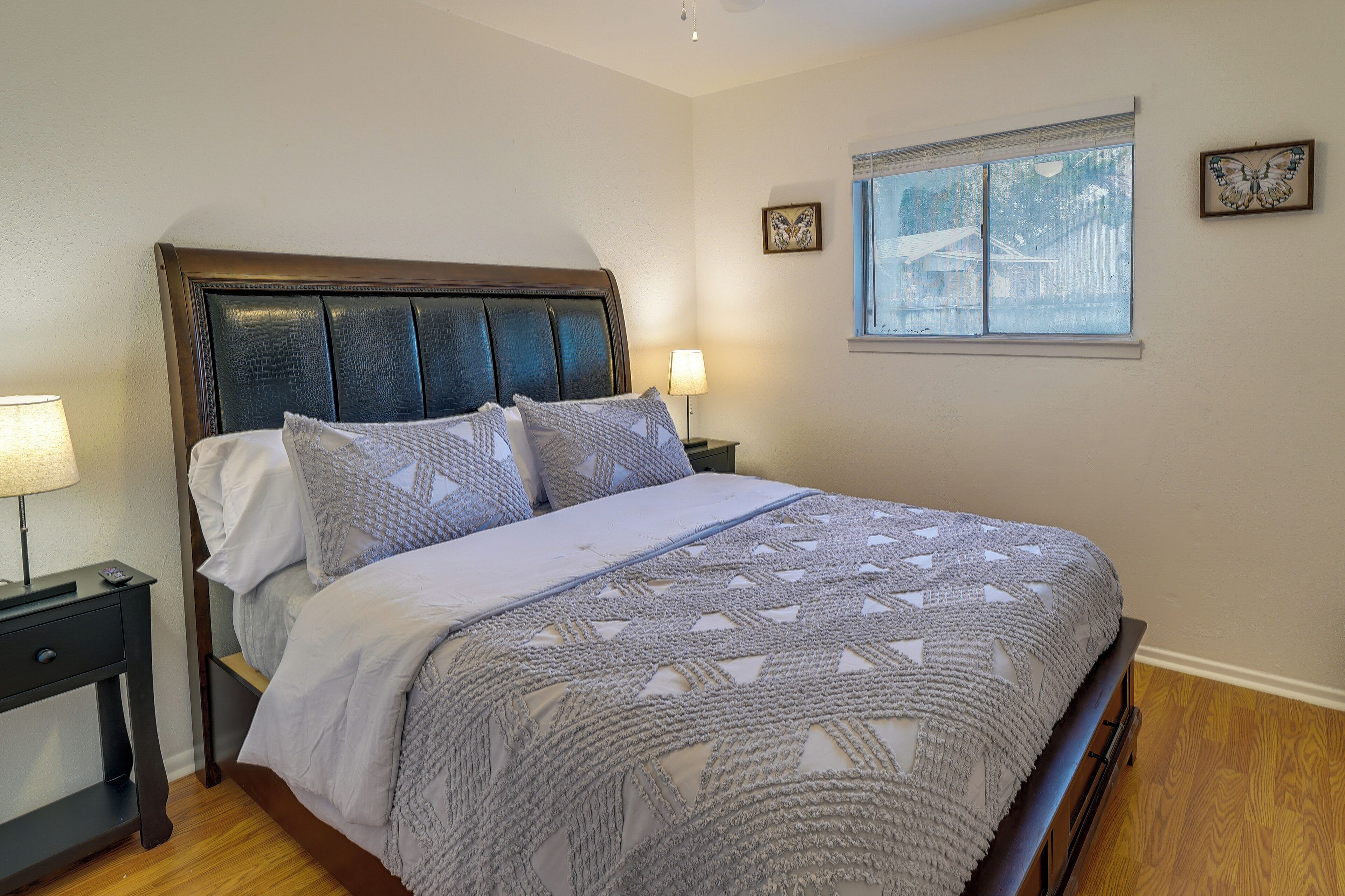 Una cama bien hecha con una colcha estampada, una lámpara de noche y una ventana con vista a una casa.