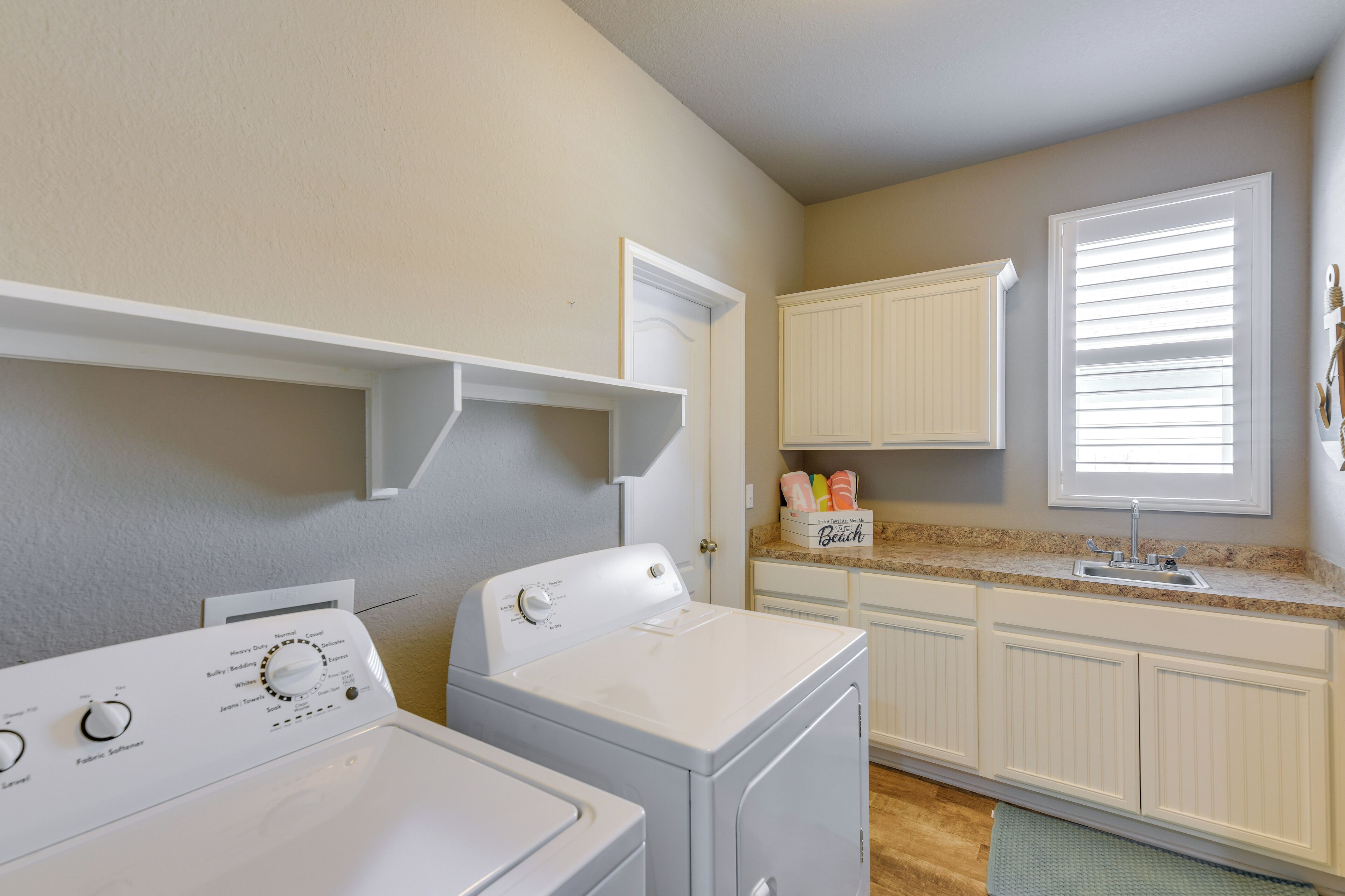 A laundry area with two white washing machines, a countertop with a sink, and a window with blinds.