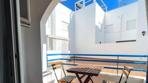 Varanda com mesa e cadeiras de madeira, parede branca e guarda-corrimão azul.