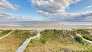 A beach with a wooden boardwalk leading to the shore, grassy dunes, and a clear sky with clouds. - Spacious Isle of Palms Home w/ Ocean Views! (Isle of Palms)