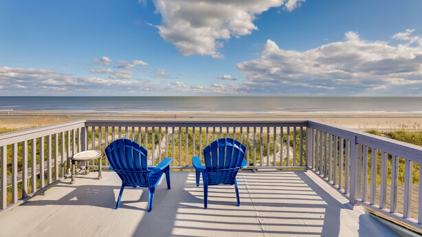 A wooden deck with two blue Adirondack chairs and a small table, overlooking a beach with a clear sky. - Spacious Isle of Palms Home w/ Ocean Views! (Isle of Palms)