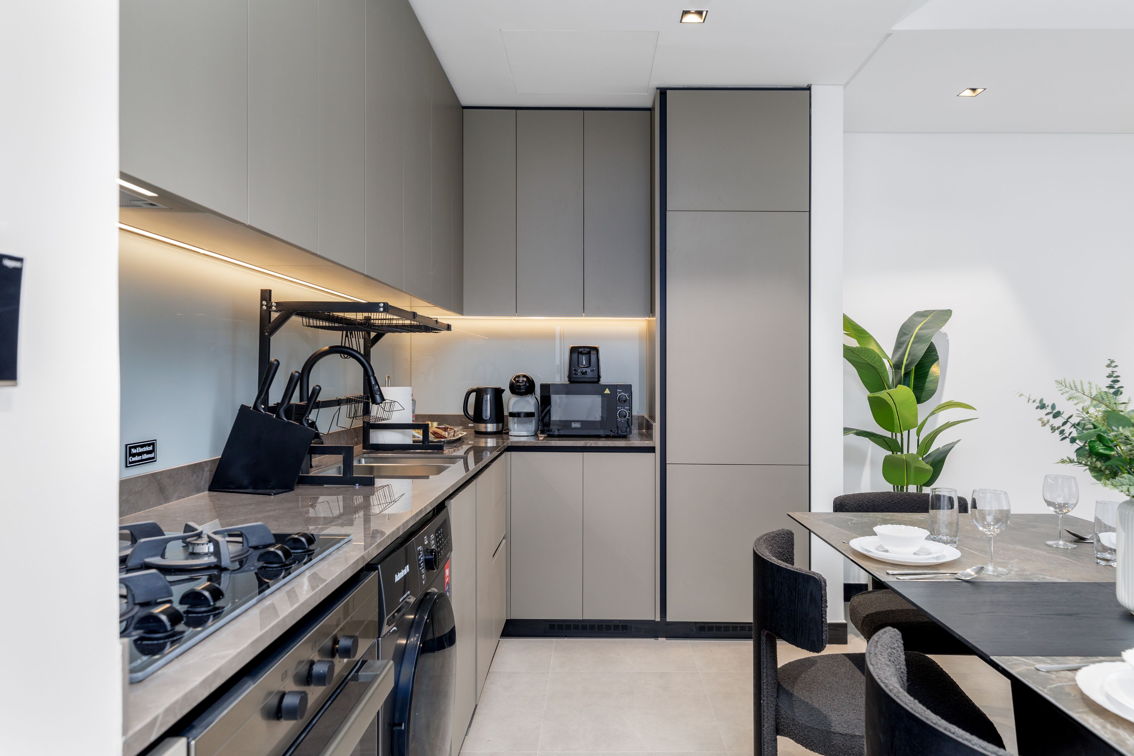 A modern kitchen with a built-in stove, microwave, and coffee maker. There is a dining table with chairs and a potted plant.