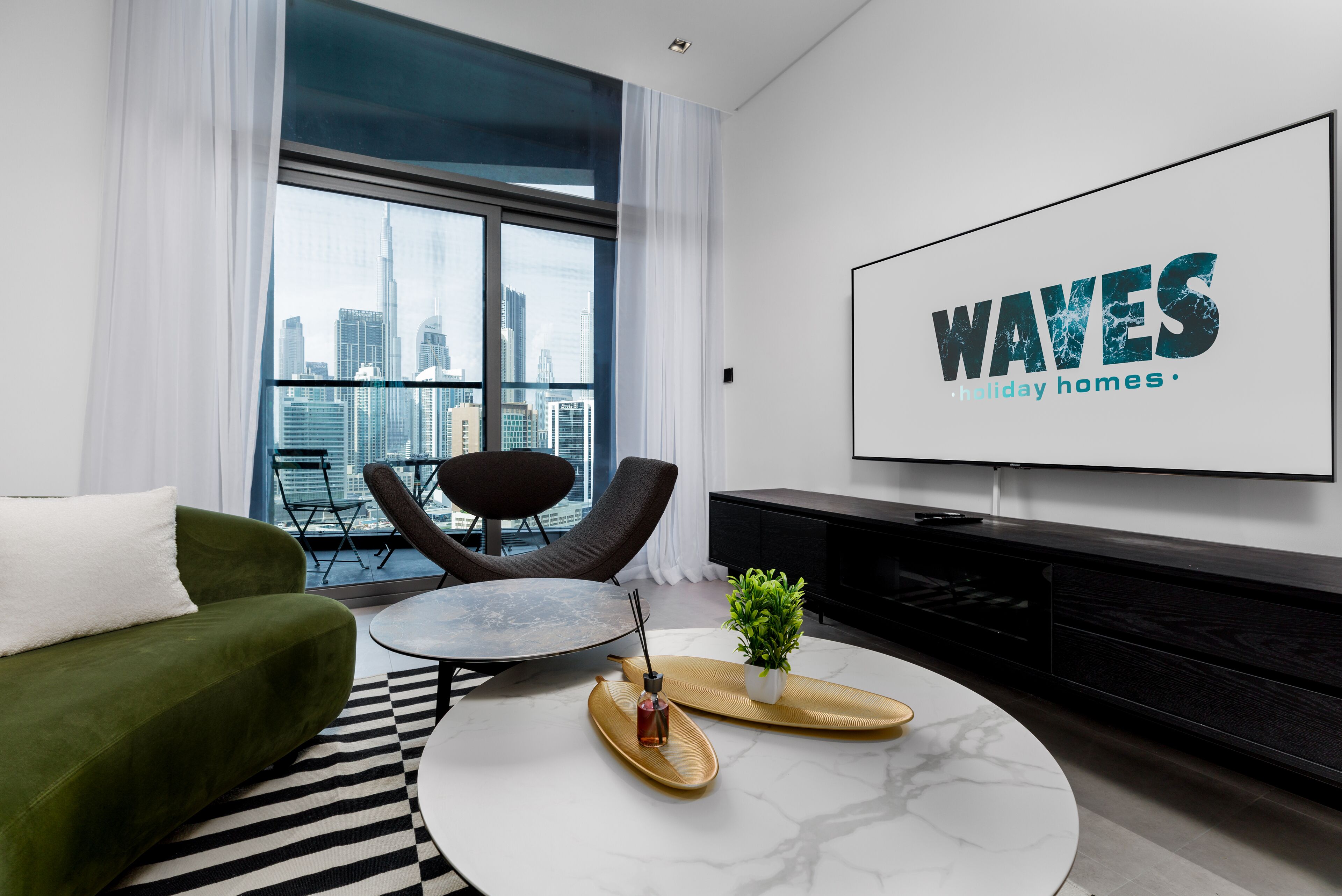 A modern living room with a green sofa, a round coffee table, a TV mounted on the wall, and a city view through large windows.