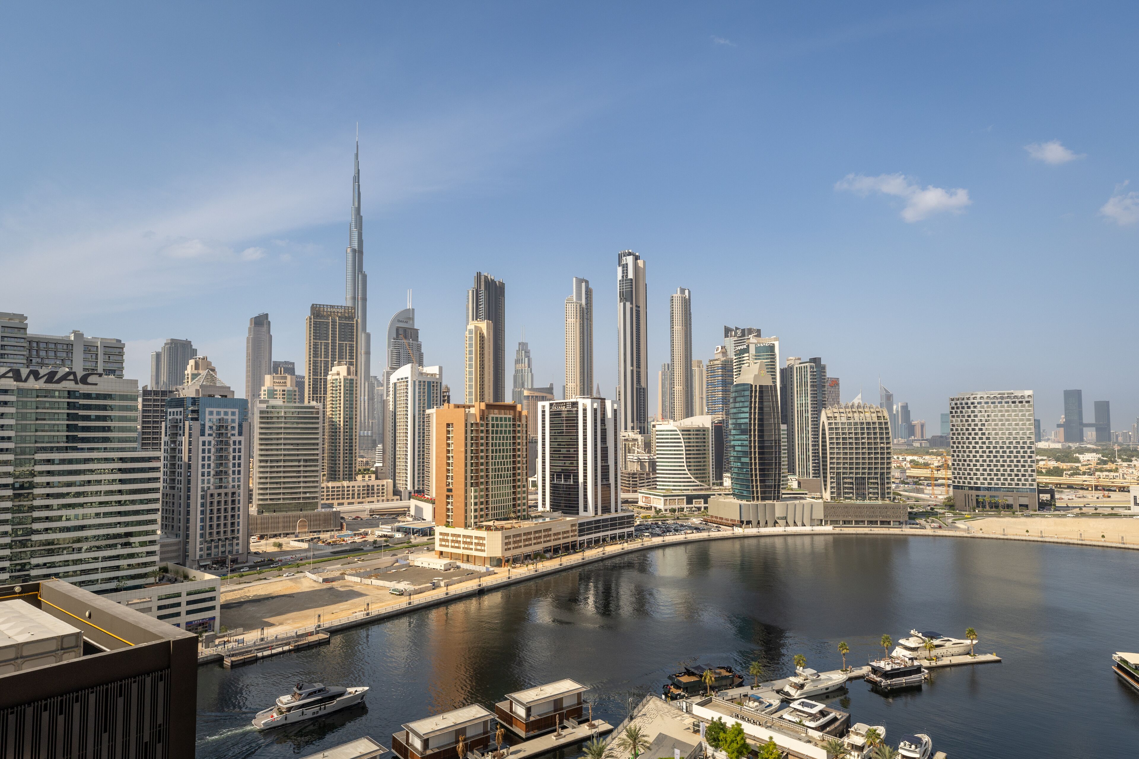 A city skyline with modern high-rise buildings, a marina with boats, and a clear sky.