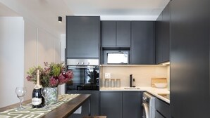 A modern kitchen with dark cabinetry, a built-in oven, microwave, and dishwasher. There is a wooden bench and a vase with flowers on the counter. - Portswood 103 (Cape Town)