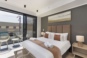 A modern hotel room with a large bed, a balcony with seating, and a city view.