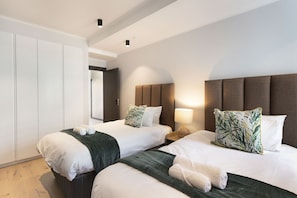 A hotel room with two beds, each with white linens and green throw blankets. There is a nightstand with a lamp and a decorative pillow. The room has a large wardrobe and a door leading to another room. - Portswood 103 (Cape Town)
