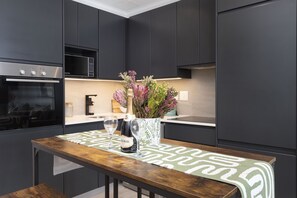 A modern kitchen with dark cabinetry, a Bosch oven, a microwave, a wooden table with a green and white patterned runner, and a vase with flowers. - Portswood 103 (Cape Town)