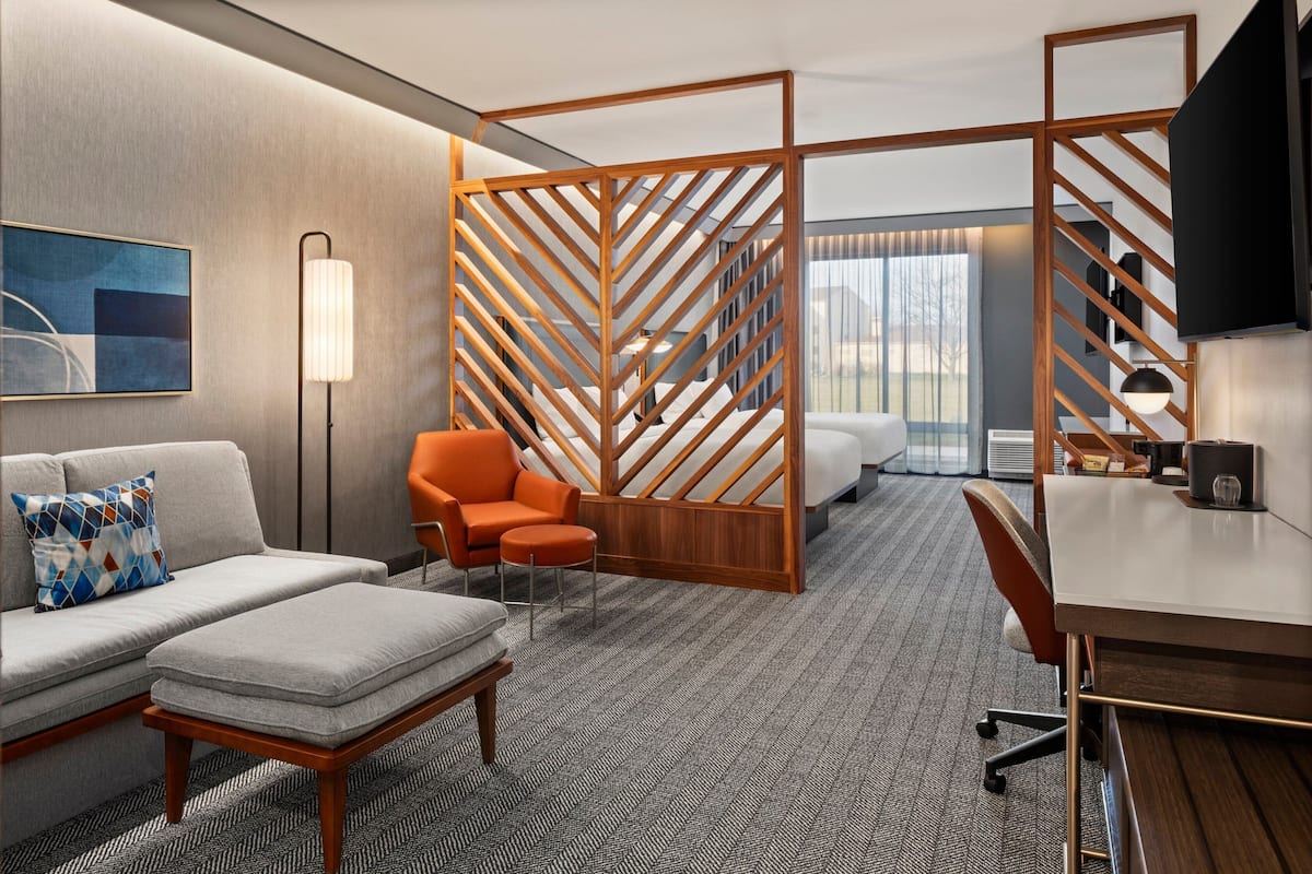 Une chambre d’hôtel équipée d’un canapé, d’un fauteuil, d’un pouf, d’un bureau et d’une grande fenêtre.