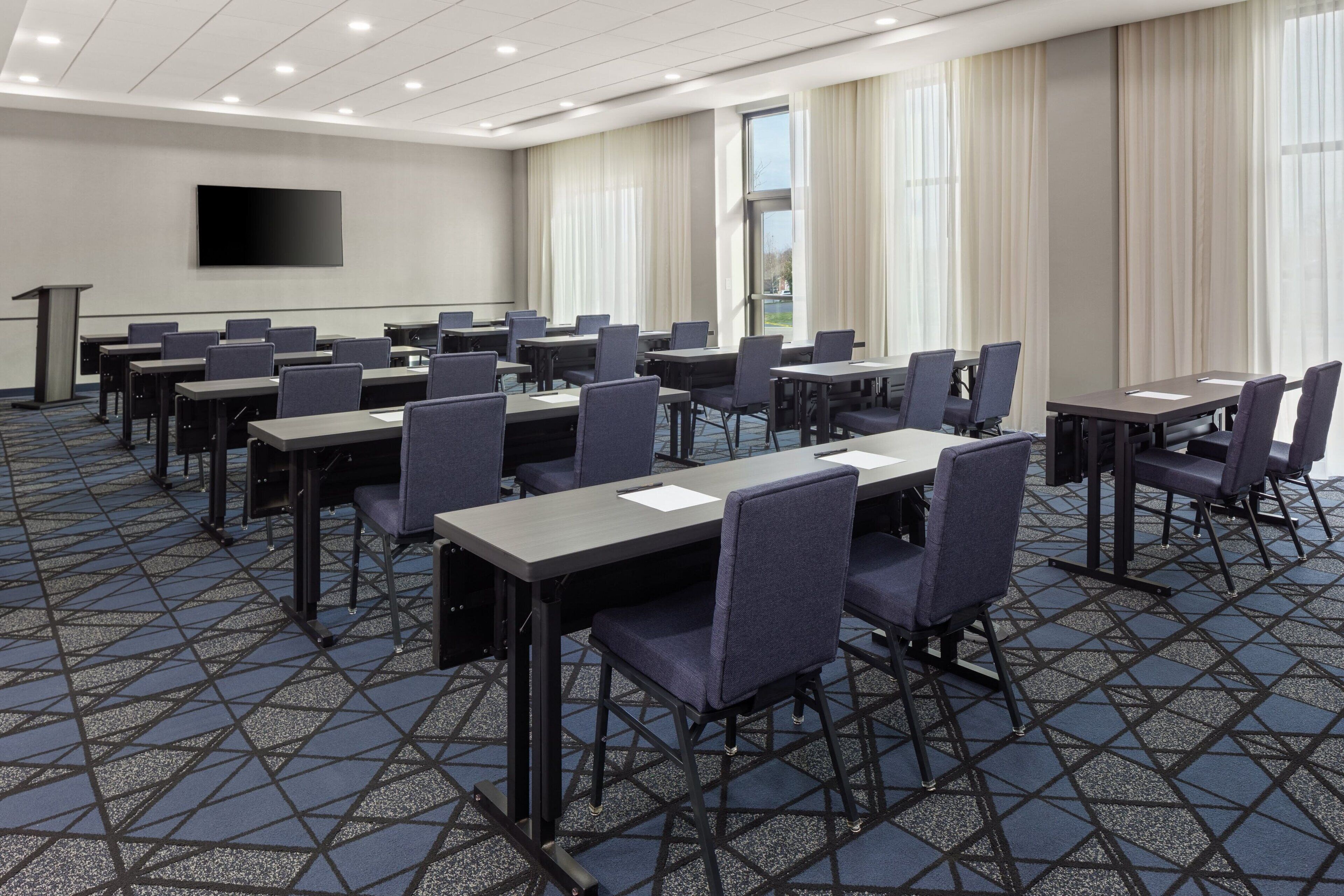 Une salle de conférence équipée de plusieurs tables et chaises, d’un écran de projection et d’un pupitre.