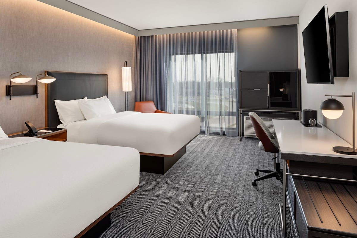 Une chambre d’hôtel avec deux lits, un bureau, une chaise, une télévision et une grande fenêtre avec des rideaux.