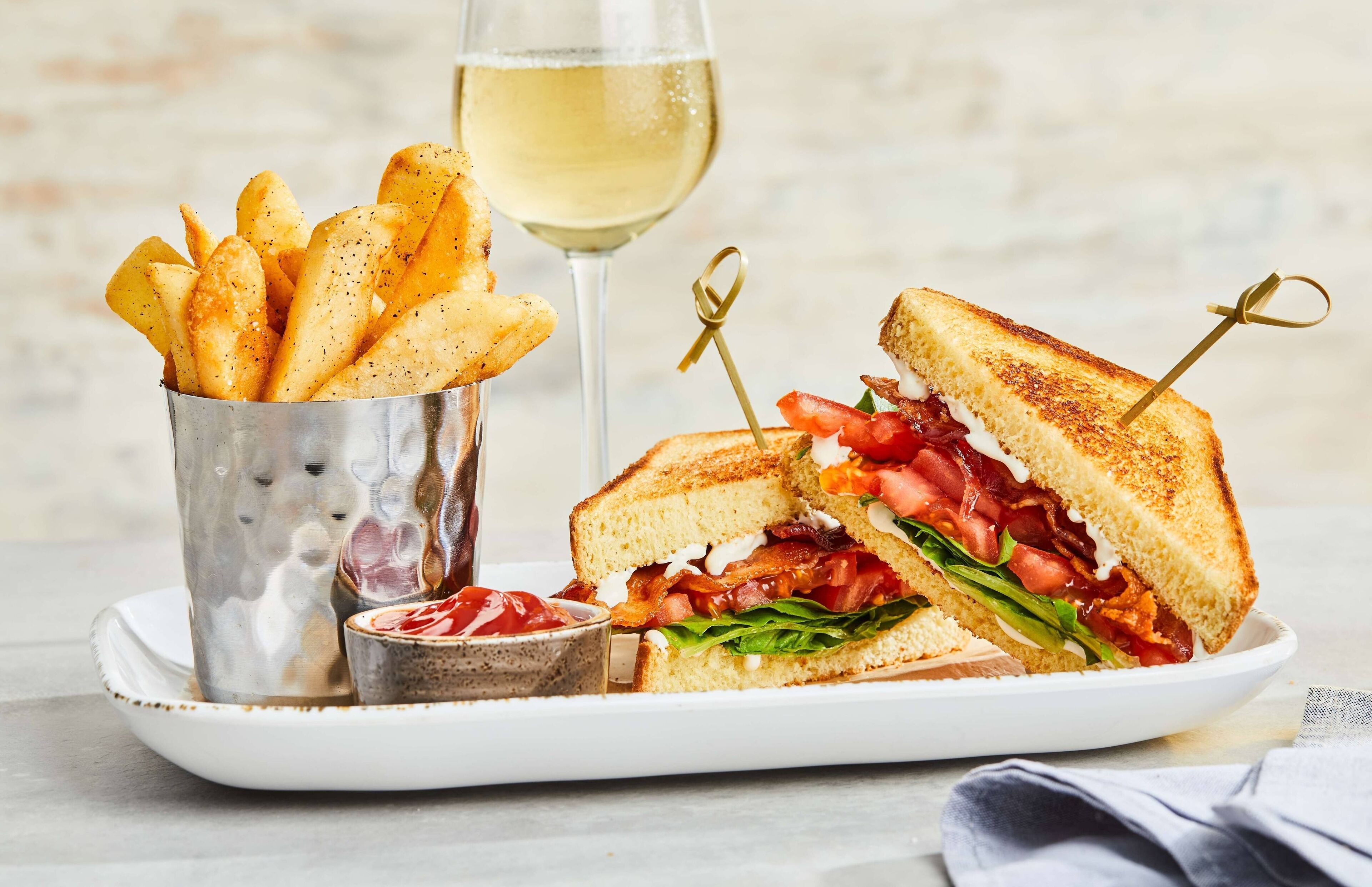A plate with a grilled sandwich, a glass of white wine, and a container of fries.