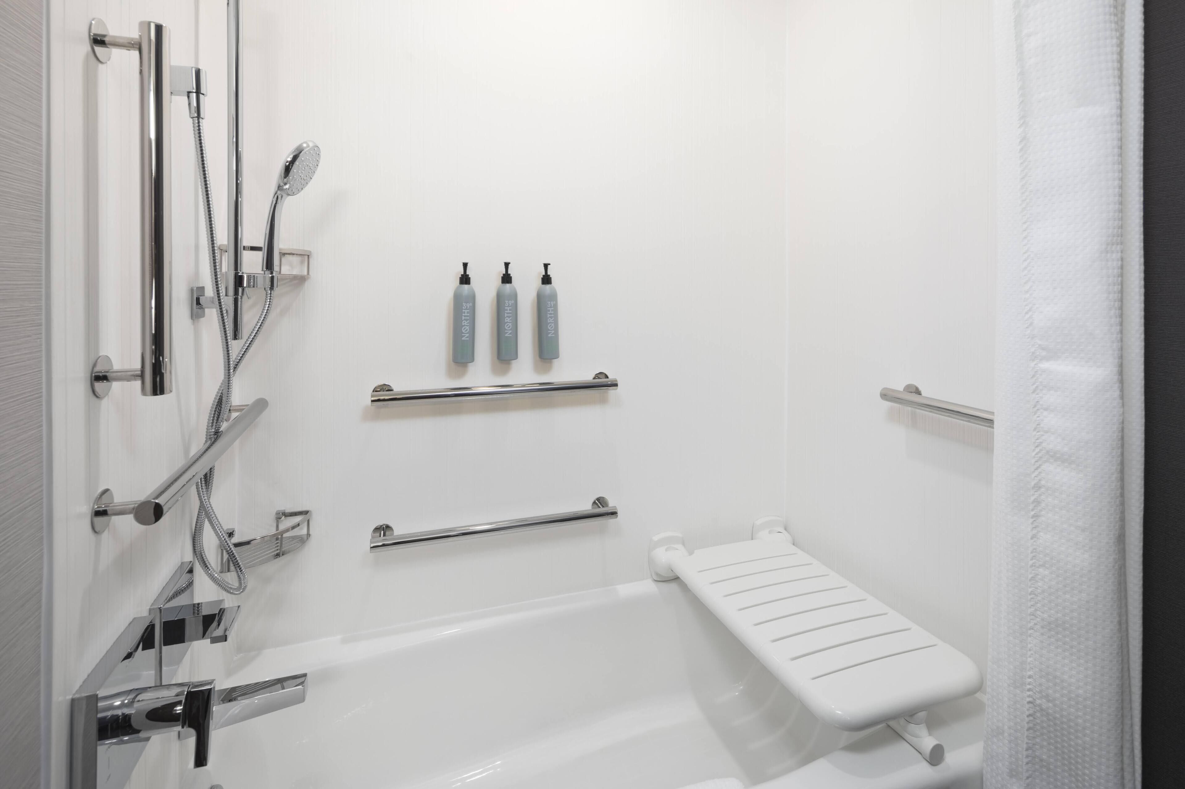 A modern bathroom with a walk-in shower, grab bars, a white bathtub, and three bottles of shampoo on a shelf.