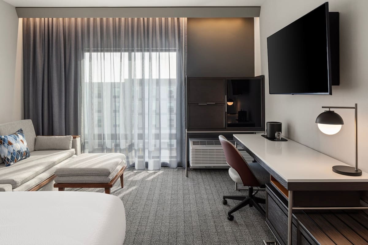 Une chambre d’hôtel avec un lit, un bureau, une chaise, une télévision et une fenêtre avec des rideaux.