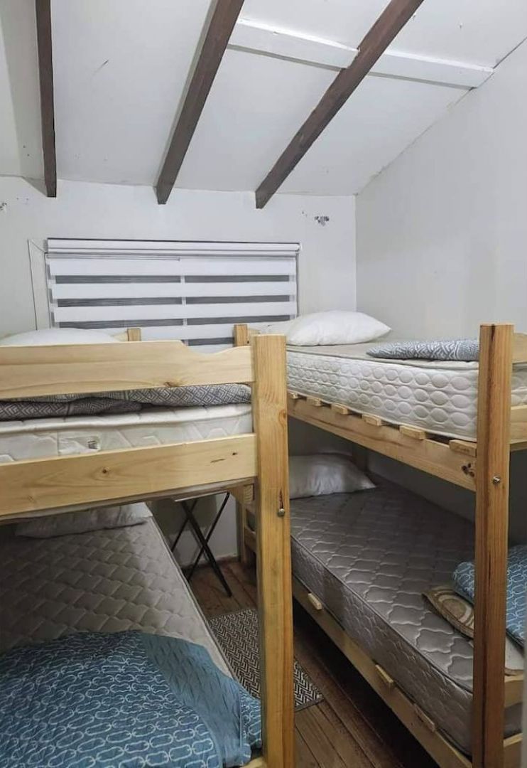 A dormitory room with bunk beds, a window with blinds, and a wooden ceiling.