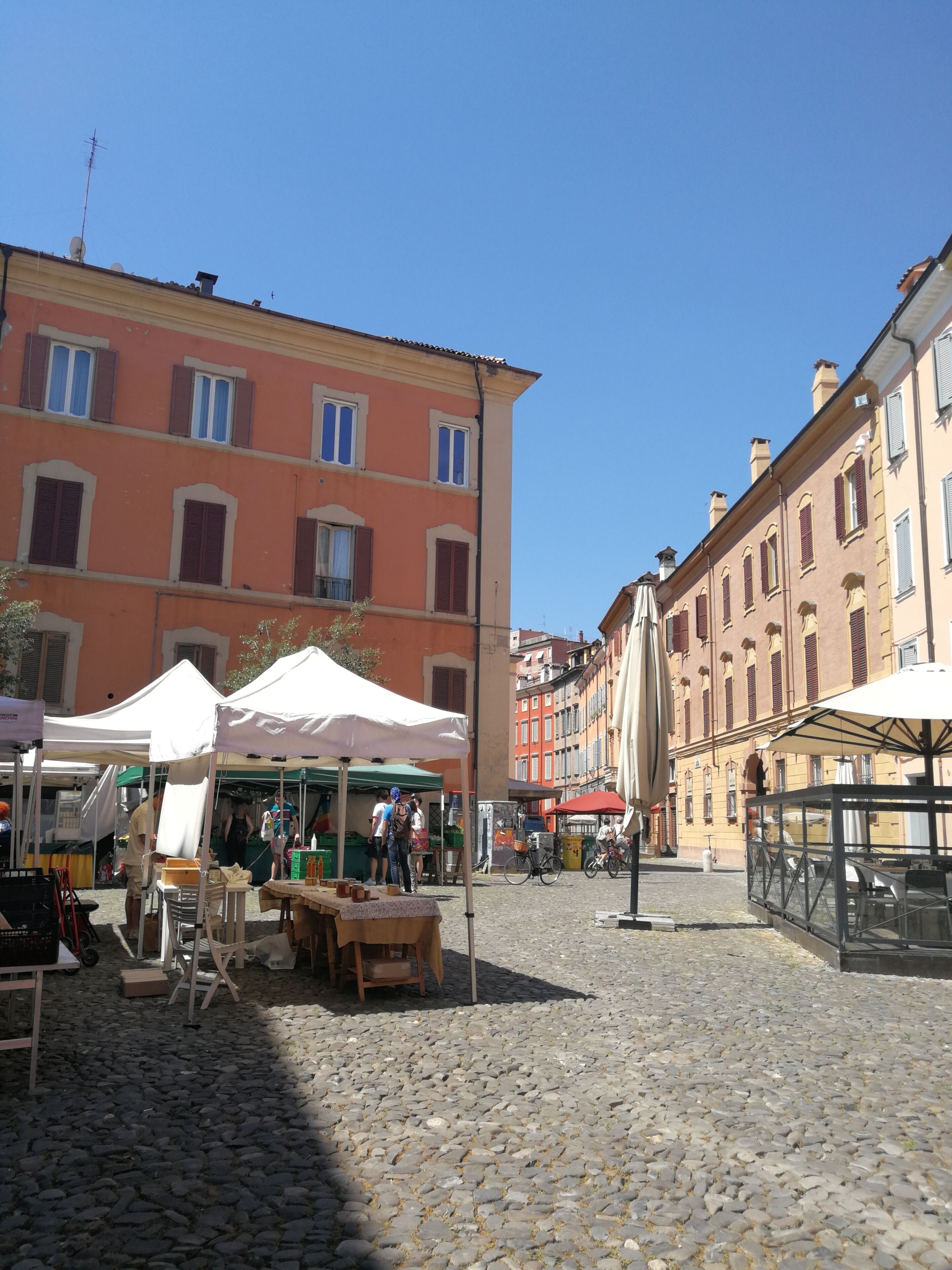 Une place pavée, bordée de bâtiments, avec des tables installées à l’extérieur et un ciel dégagé.