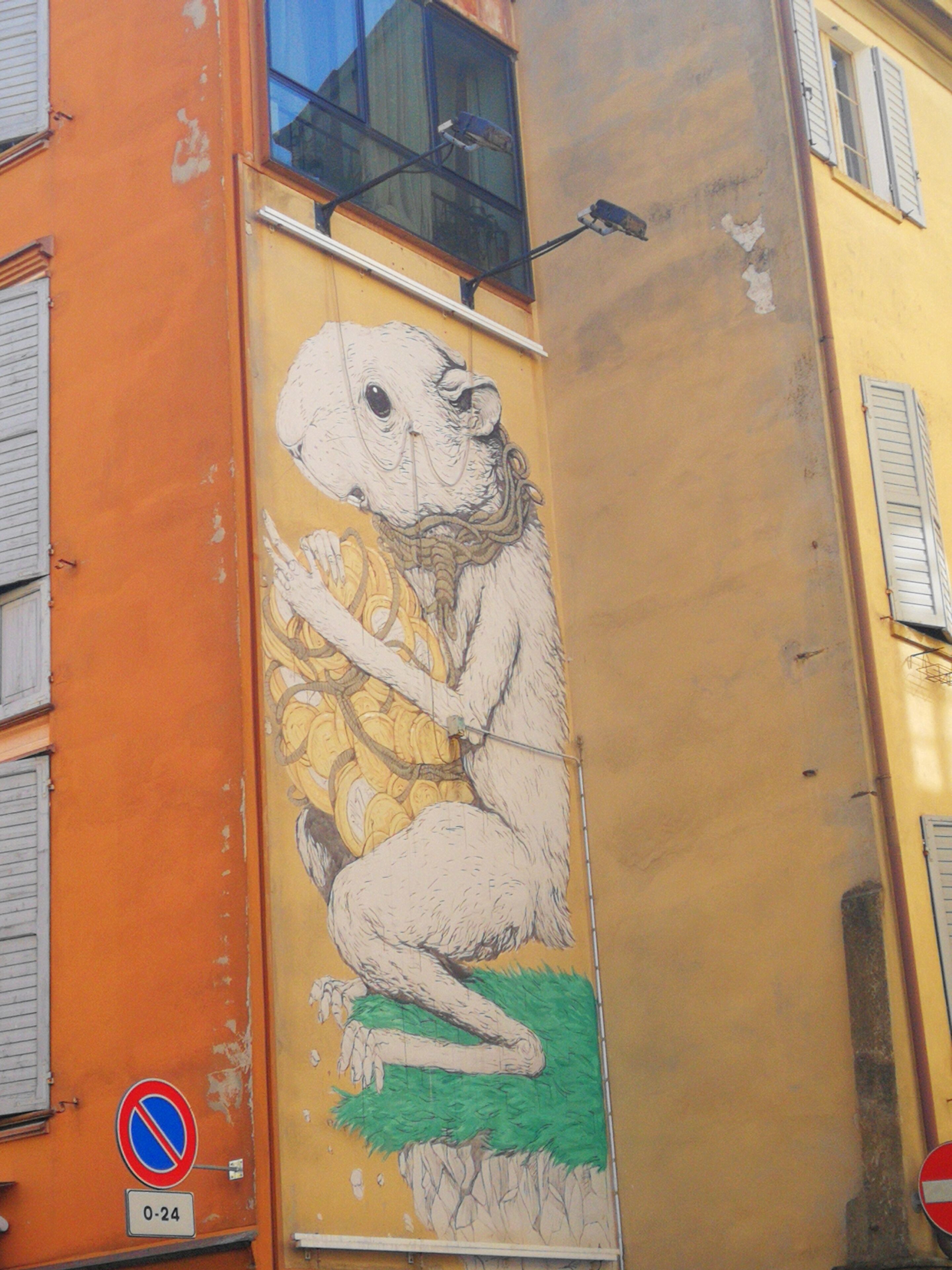 Un bâtiment orné d’une fresque représentant une créature tenant des pièces de monnaie, d’un panneau indiquant qu’il est interdit de se garer et d’un panneau indiquant « 0-24 ».