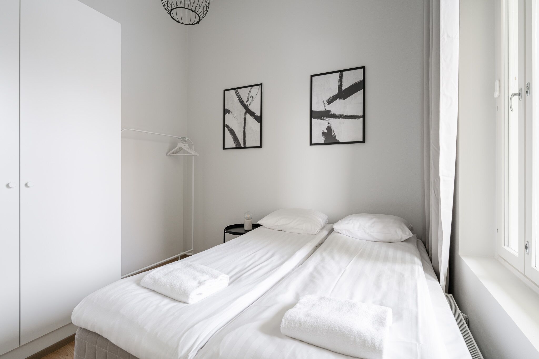 Ein schlicht eingerichtetes Schlafzimmer mit einem Bett, weißen Vorhängen und zwei abstrakten Bildern an der Wand.