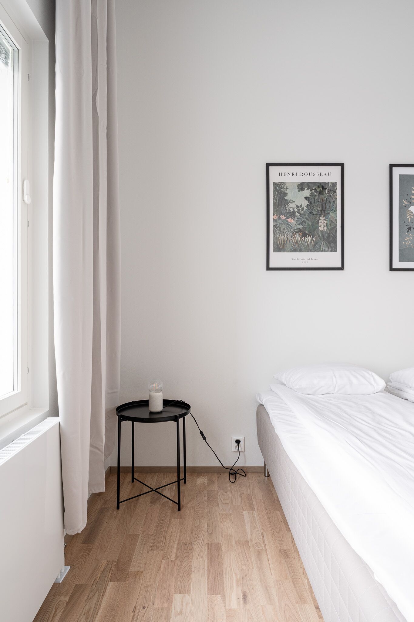 Ein Schlafzimmer mit Holzboden, weißer Bettwäsche, einem kleinen schwarzen Tisch mit Kerze und zwei gerahmten Pflanzenbildern an der Wand.