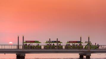 Un puente con sombrillas verdes y zonas de estar, con vista a un cuerpo de agua al atardecer.