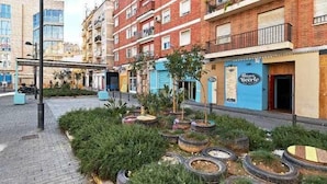 En gade med en række bygninger, hvoraf den ene har et skilt med 'Illegible Beer' og en anden en blå markise. Der er plantekasser med grønne planter og dæk brugt som plantekasser.