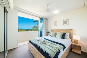 A hotel room with a large bed, a nightstand, a window with a view of the sea, and a ceiling fan.