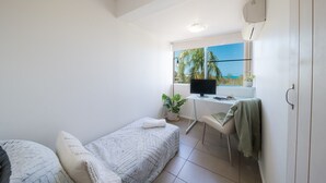 A compact room with a bed, a desk with a computer, a window with a view of palm trees, and a small plant. - Picture Perfect On Azure (Airlie Beach)