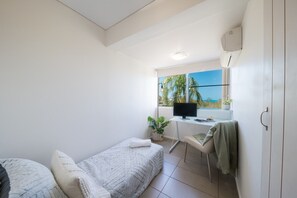 A compact room with a bed, a desk with a computer, a window with a view of palm trees, and a small plant. - Picture Perfect On Azure (Airlie Beach)