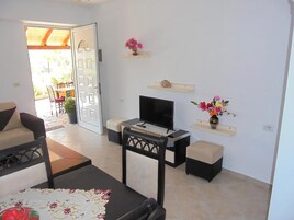 Una sala de estar con un sofá, una mesa de centro, un televisor y una pequeña mesa con un mueble para televisión. Hay jarrones con flores en la pared y una puerta que da al exterior.