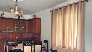 A living room with a sofa, coffee table, dining table with chairs, and a kitchen area with cabinets and a chandelier.