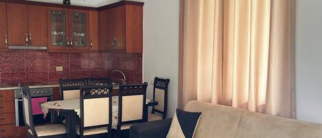 Una sala de estar con sofá, mesa de centro, comedor con sillas y una zona de cocina con armarios y una araña.
