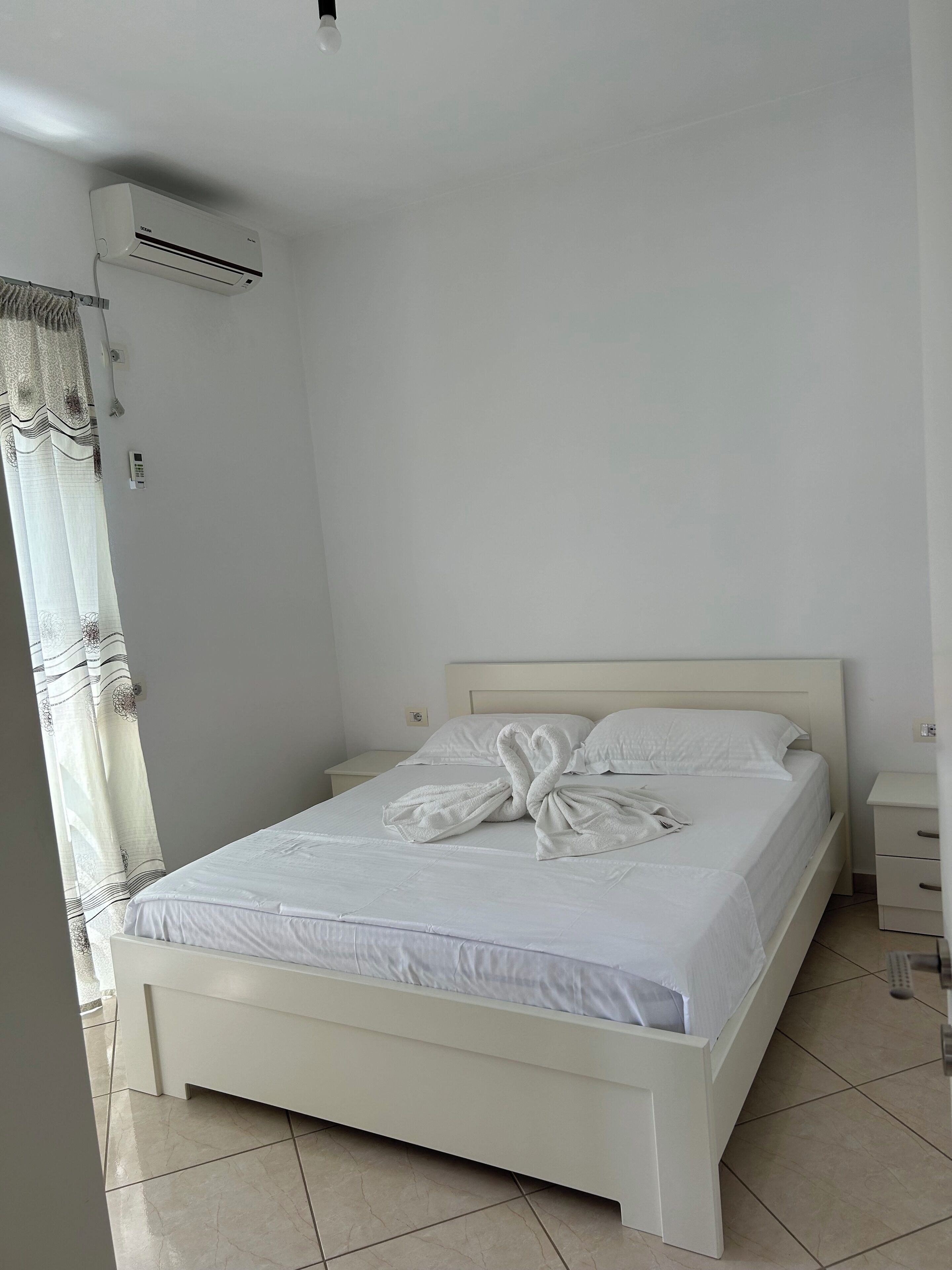A neatly made bed with a towel in the shape of a swan, a white bedside table, a nightstand, a wall-mounted air conditioner, and a curtain with a pattern.