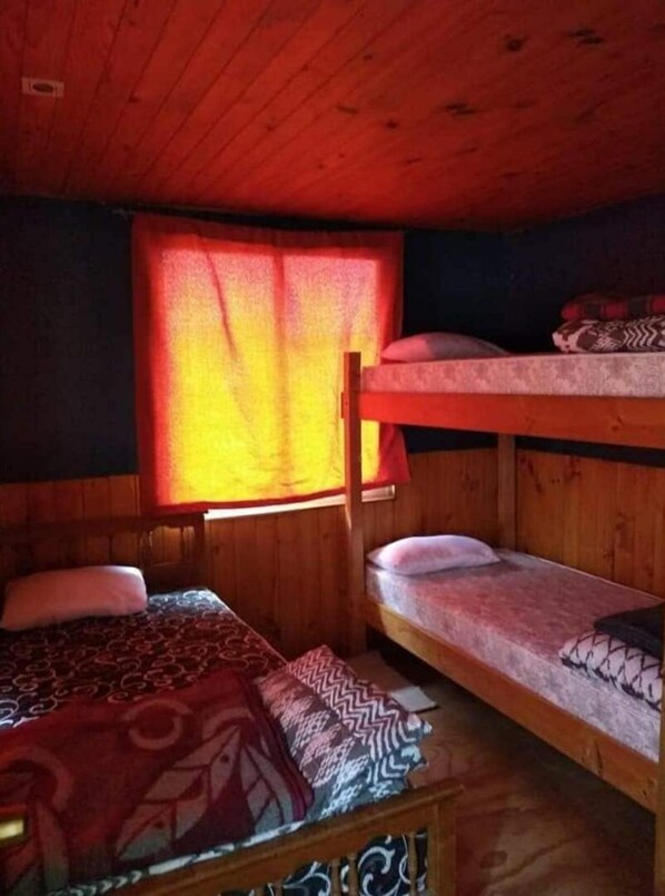 Habitación con literas, techo y paredes de madera, una ventana con cortinas rojas y una colcha estampada.