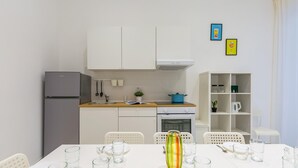 A modern kitchen with white cabinetry, a grey refrigerator, a blue pot on the counter, and a dining area with a white table set for two. - White Blue Apartment at Sanit by Wonderful Italy (Naples)