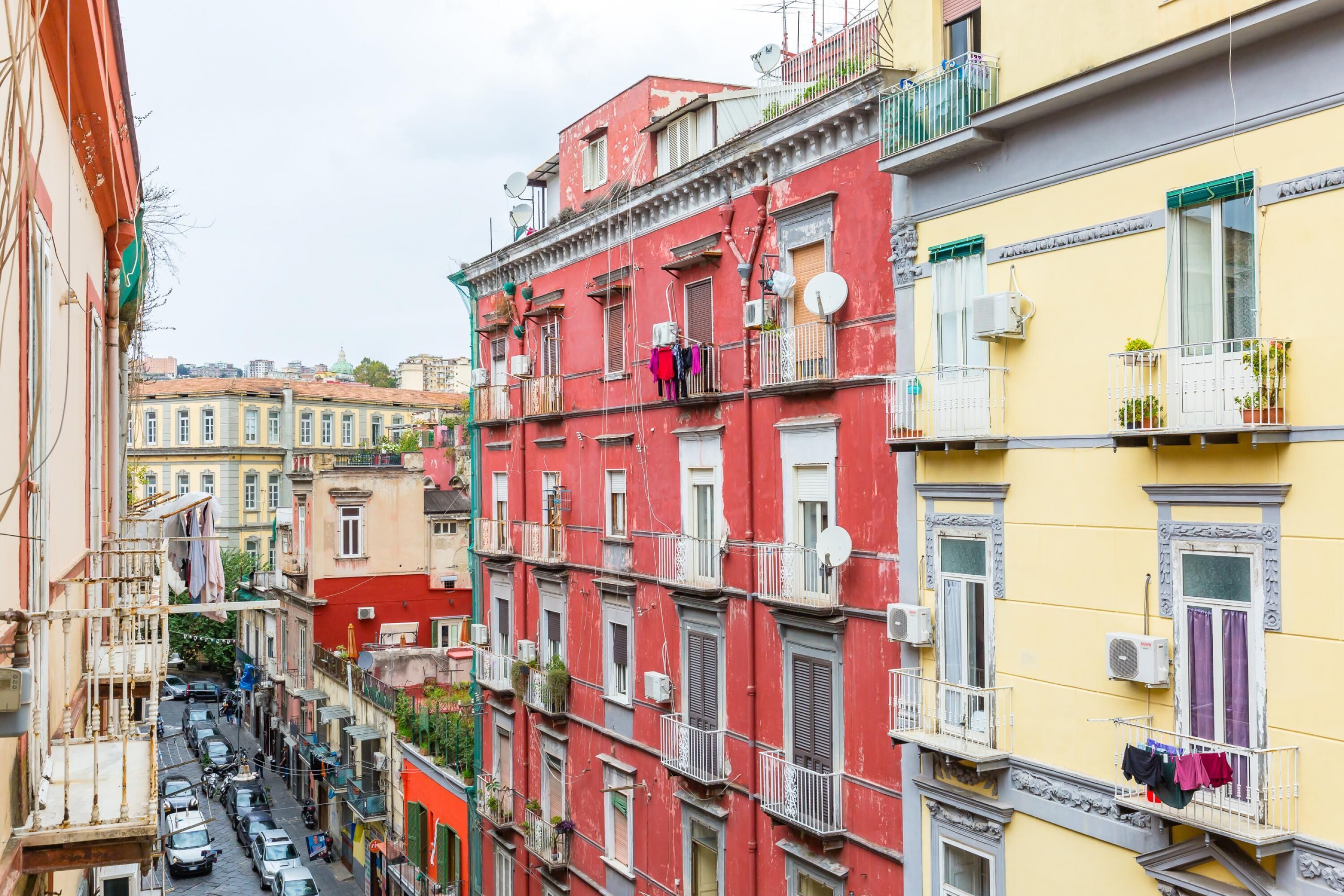 A narrow street lined with multi-story buildings, balconies with clothes hanging, and parked cars.