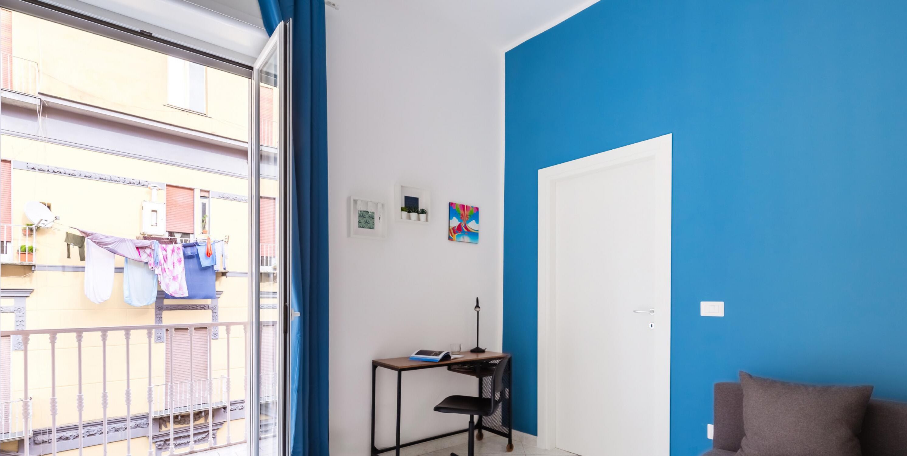 Une pièce avec un mur d’accent bleu, une porte, une petite table avec une lampe et une vue sur un balcon où du linge est suspendu pour sécher.