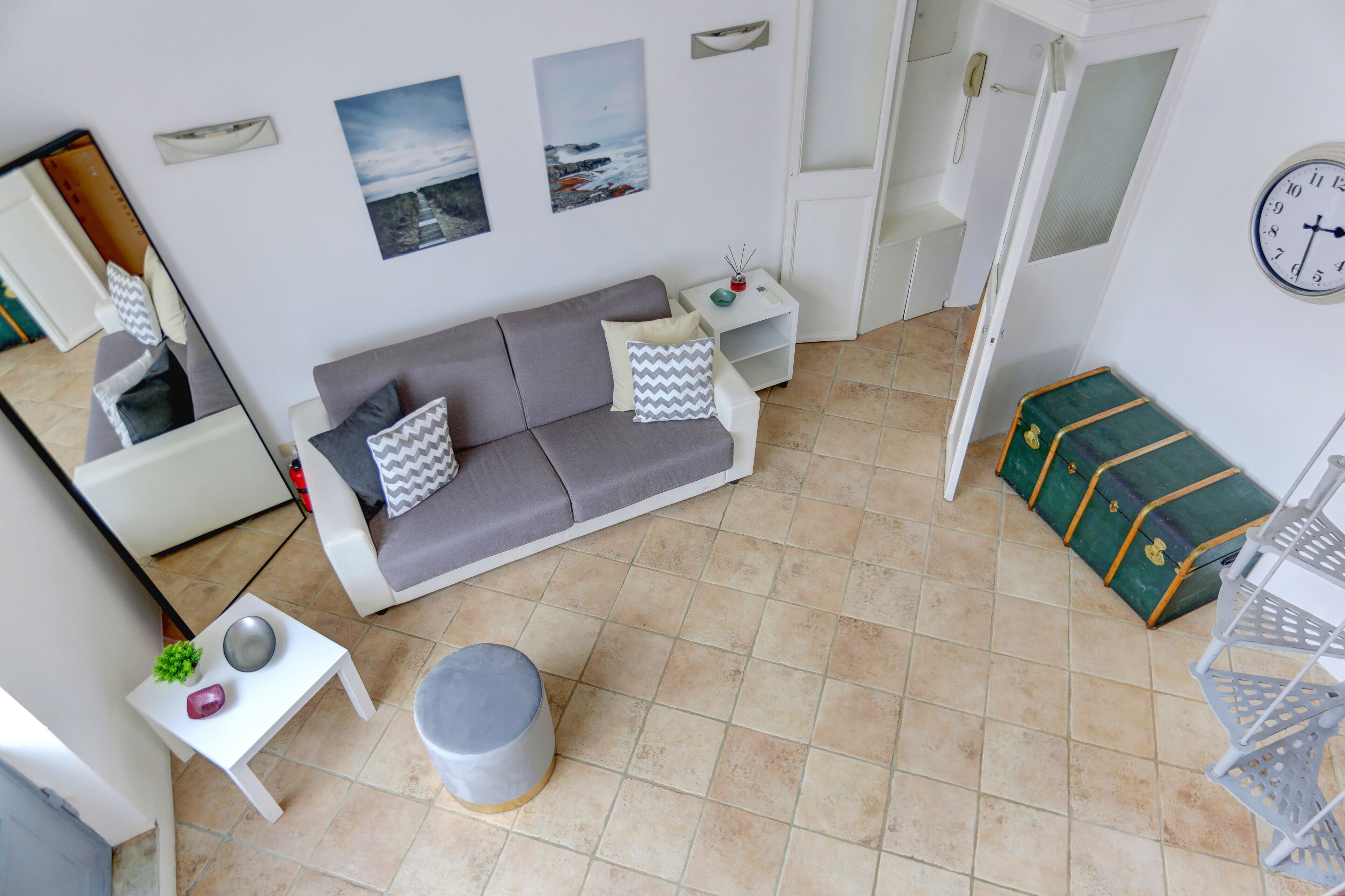 Ein modernes Wohnzimmer mit einem grauen Sofa, weißen Beistelltischen, einem runden Hocker und einem grünen Koffer.