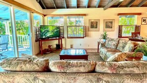 A living room with a large flat-screen TV, a sofa set, a coffee table, and a large window offering a view of greenery.