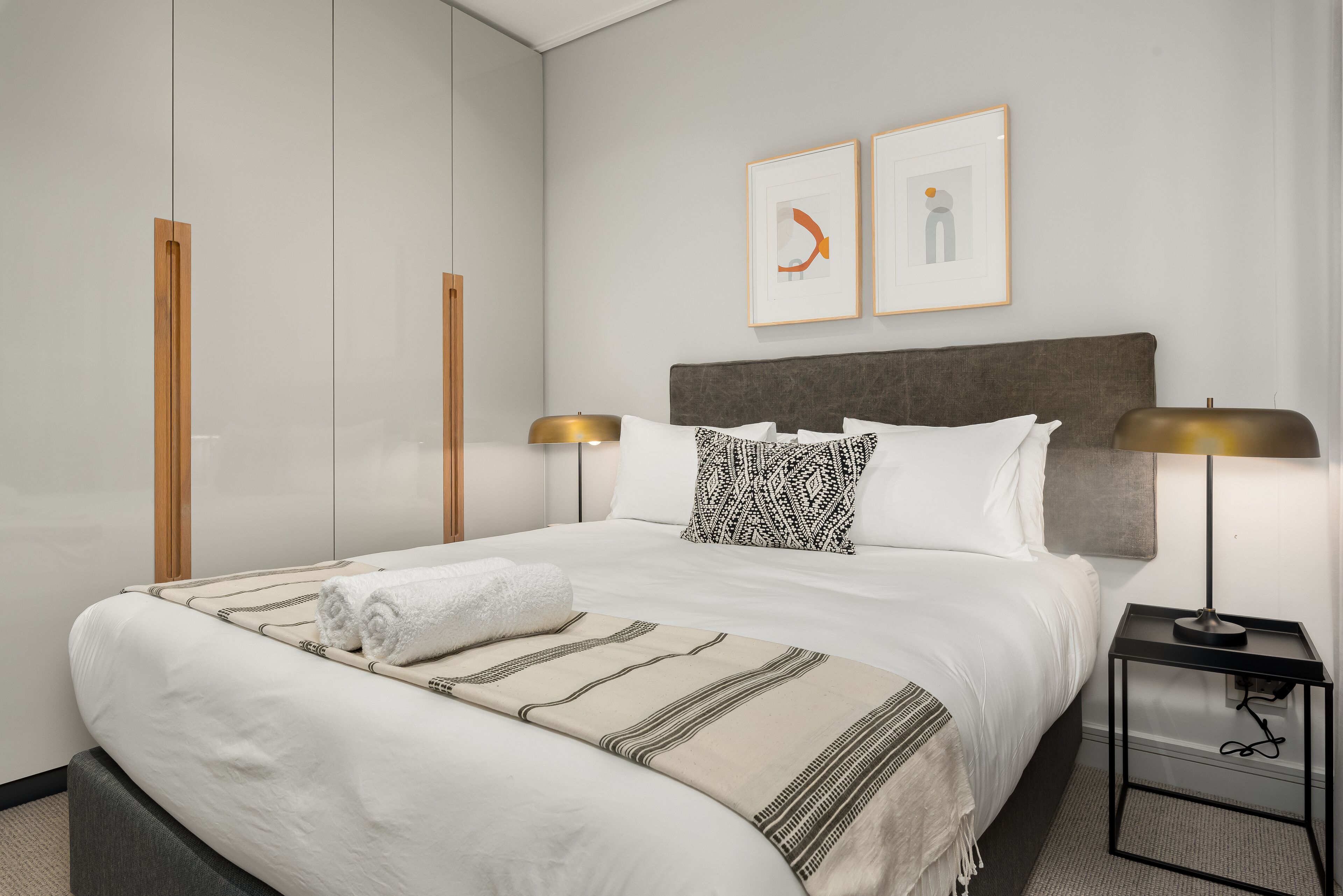 A neatly made bed with a white comforter, a beige throw blanket, and a rolled white towel. There are two framed artworks on the wall above the bed.