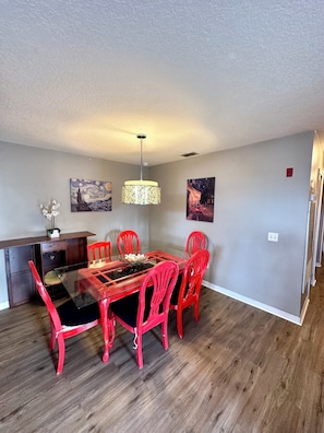 A dining area with a glass table, red chairs, a chandelier, and two paintings on the wall.