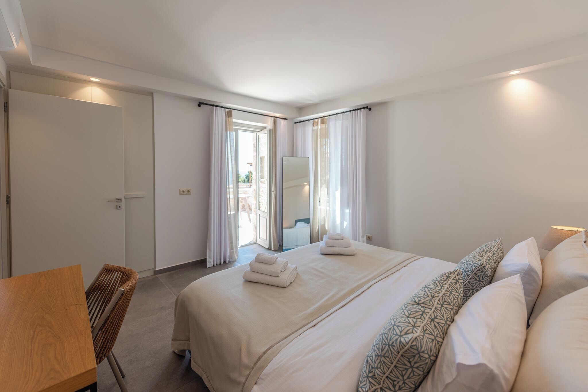 Ein Hotelzimmer mit einem großen Bett, einem Holzstuhl, einem Schreibtisch und Blick ins Freie durch das Fenster.