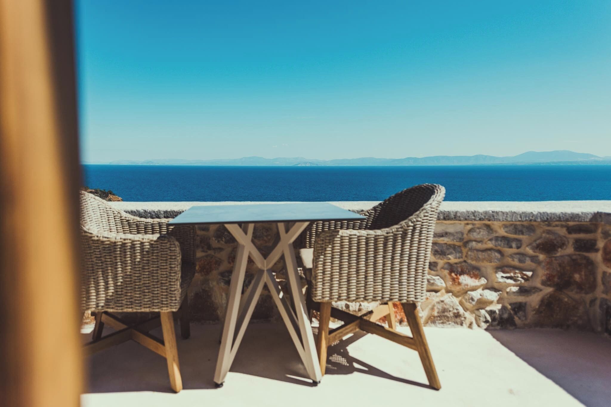 A table and two wicker chairs on a balcony overlooking the sea.