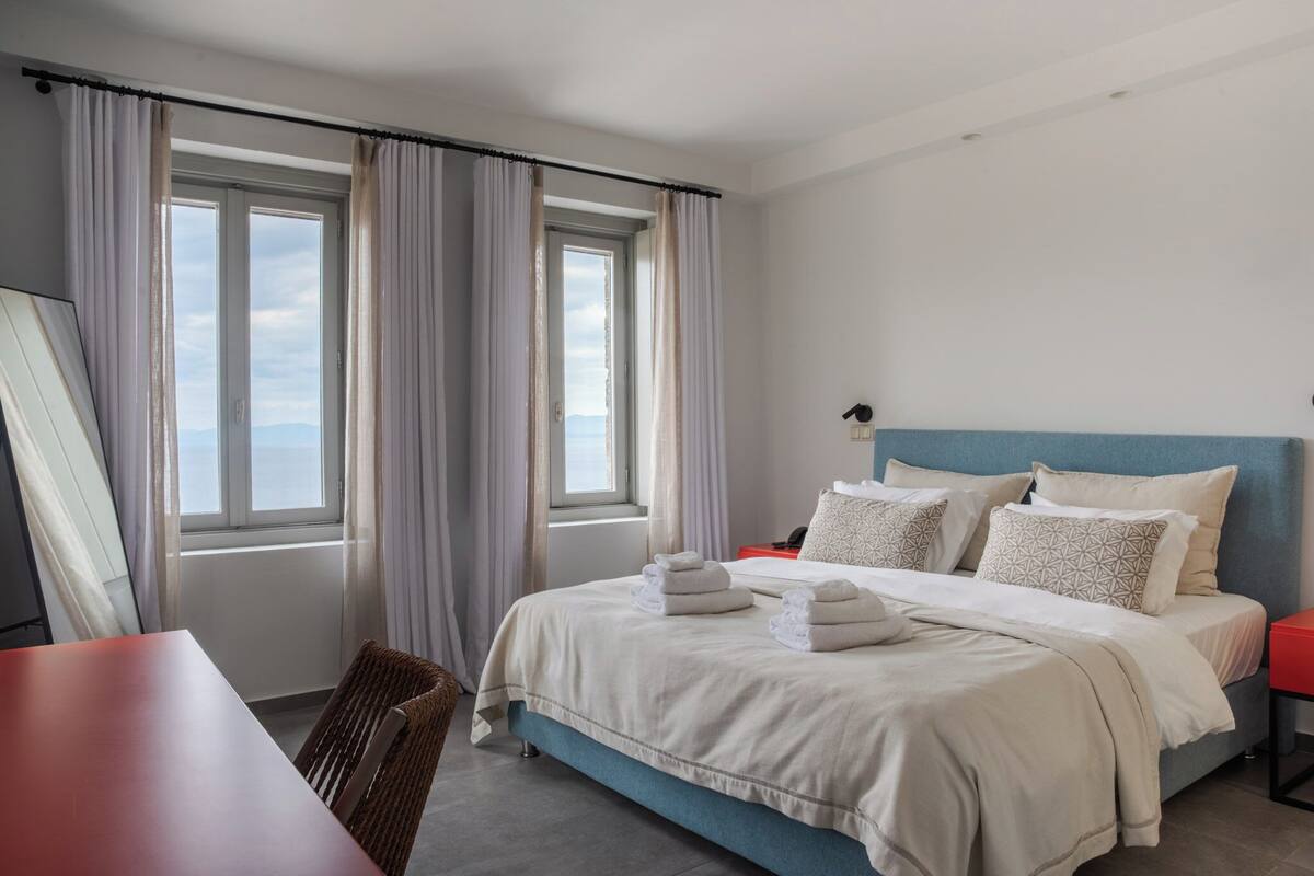 A hotel room with a bed, a table, a chair, and a view of the sky through the window.