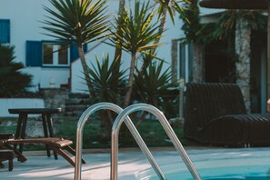 A swimming pool with metal ladders, a wooden table, and a covered wicker chair.