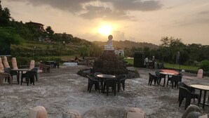 An outdoor seating area with stone tables and chairs, a central stone structure, and a view of the sunset over a hilly landscape. - Rio Forest Resort (Karjat)