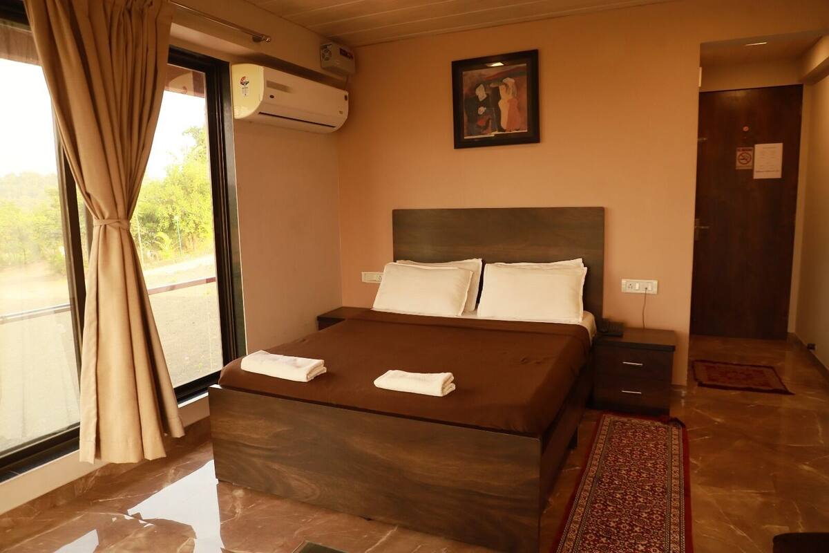 A hotel room with a bed, a window with curtains, an air conditioning unit, a painting on the wall, and a door with a sign.