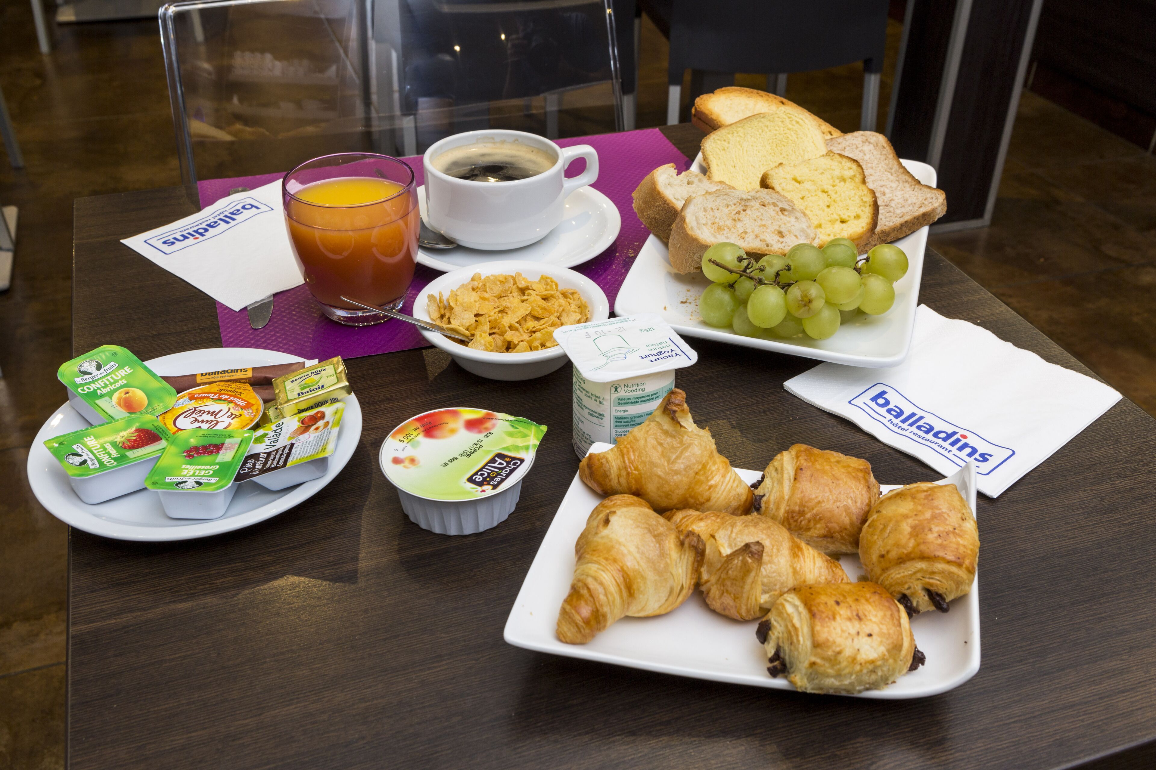 Een ontbijttafel met croissants, brood, druiven, ontbijtgranen, yoghurt en een kop koffie.