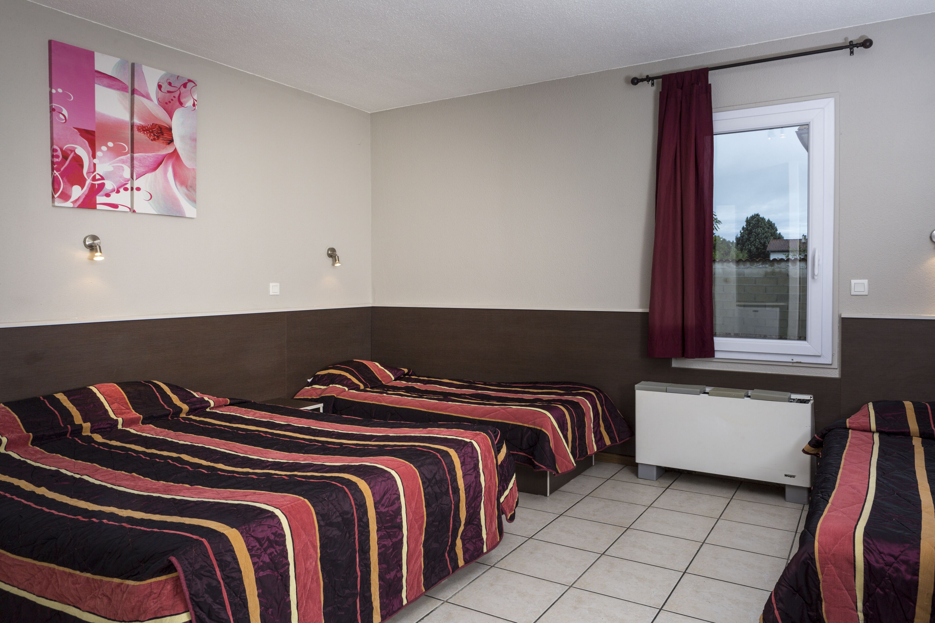 A room with three beds, a radiator, a window with curtains, and a wall-mounted light fixture.